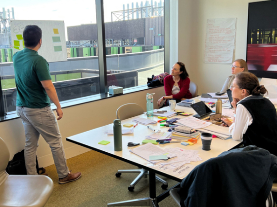Photo of Vax4Health team having a meeting. One person pointing at a work sheet, while three people sitting around a table look on.